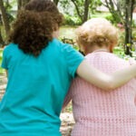 Eine junge Frau unterstützt eine ältere Dame beim Spaziergang durch einen blühenden Garten.
