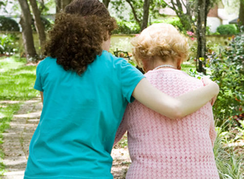 Eine junge Frau unterstützt eine ältere Dame beim Spaziergang durch einen blühenden Garten.